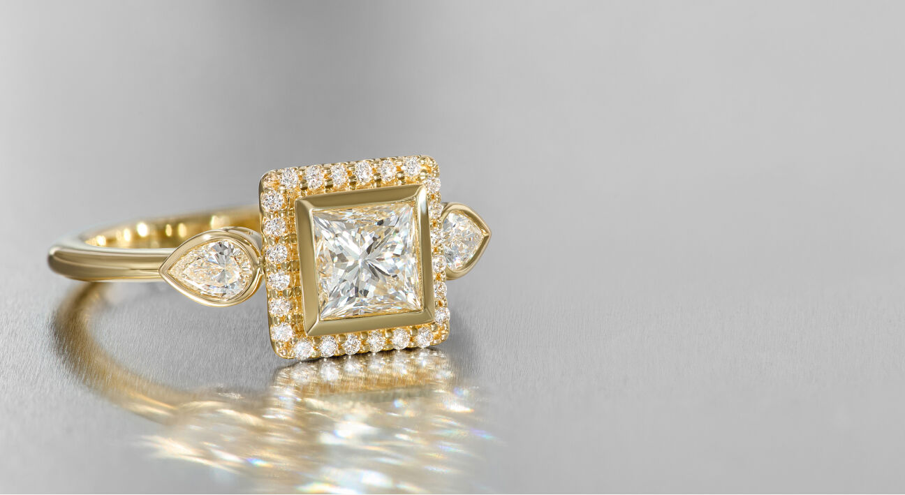 A classic diamond and yellow gold engagement ring with a princess cut centerpiece surrounded by round diamonds. The ring is reflected from a metallic surface.